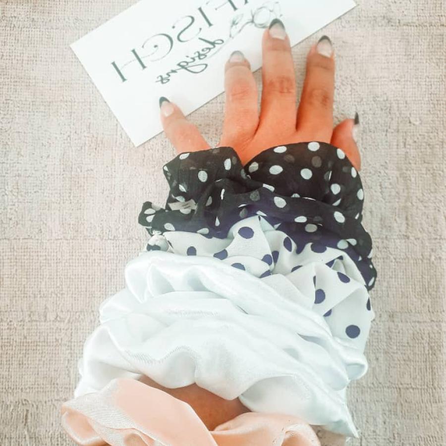 A hand with cream, white, white with black polka dots, black with white polka dots scrunchies touching a black DaFisch logo on a white envelope sized paper. 