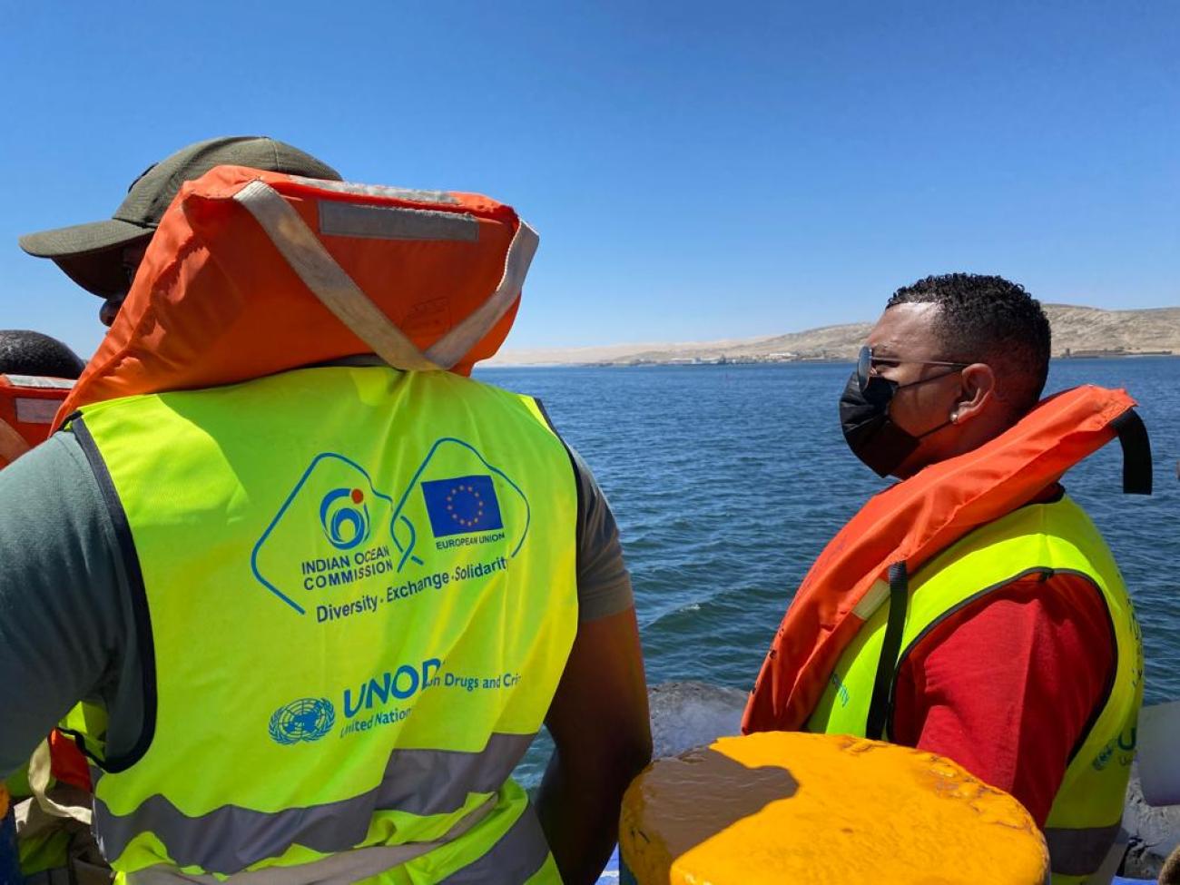 Image shows two men in hi-vis and life jackets on a boat looking out to sea