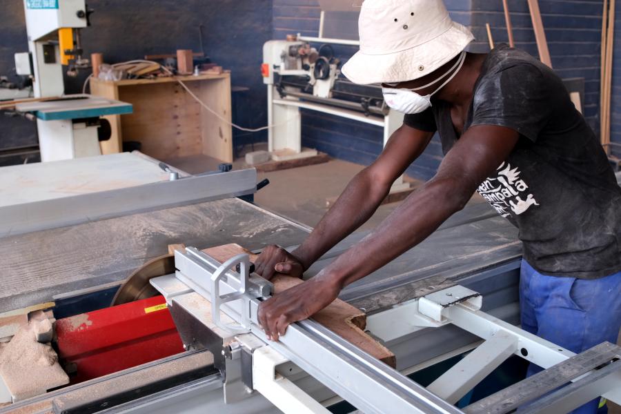 Tabled Namibia employee wearing overalls and a black shirt, cuts wood while wearing a mask.